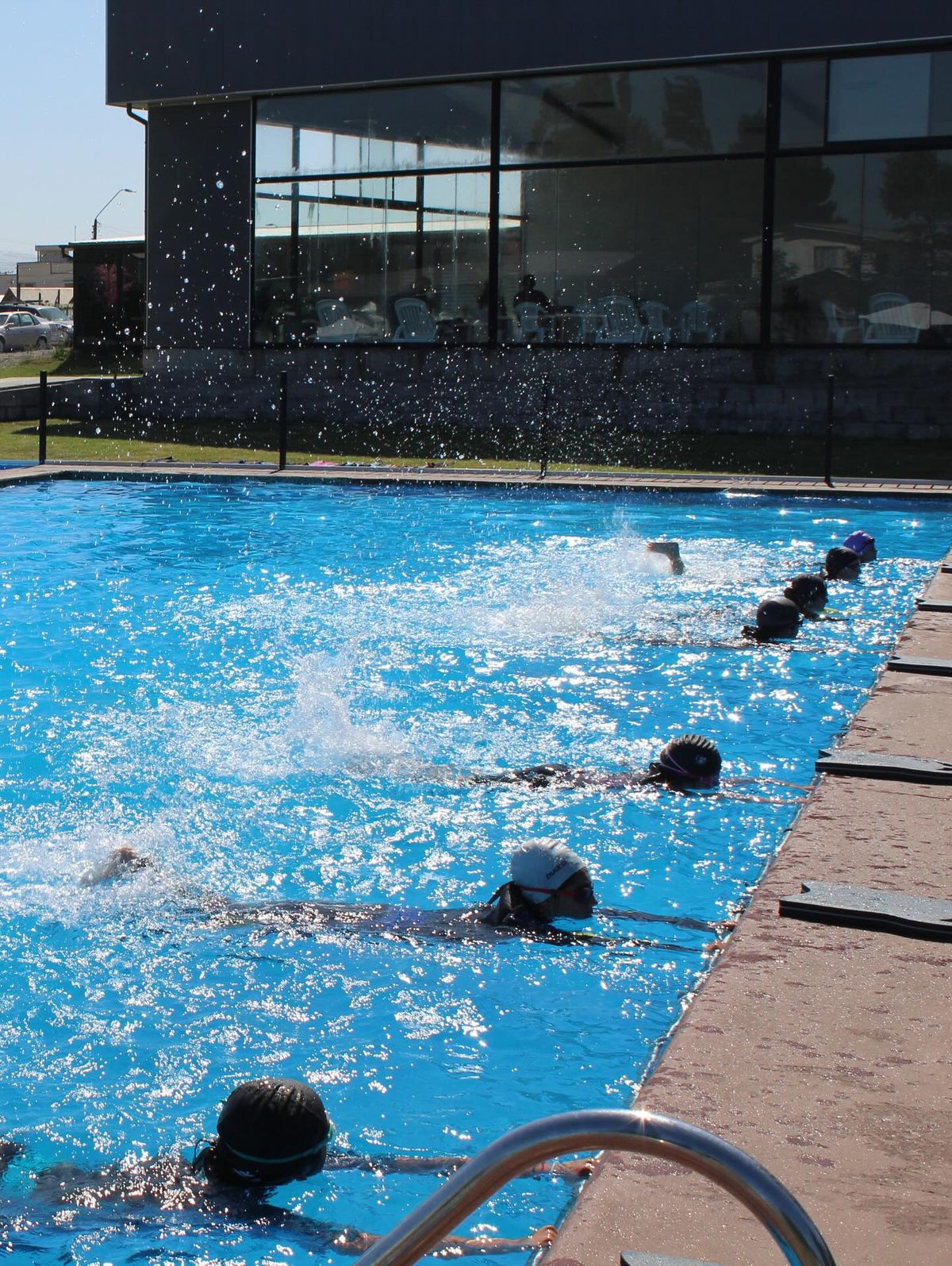 Alumnos con gorro y antiparras practicando patada tomados del borde de la piscina al aire libre