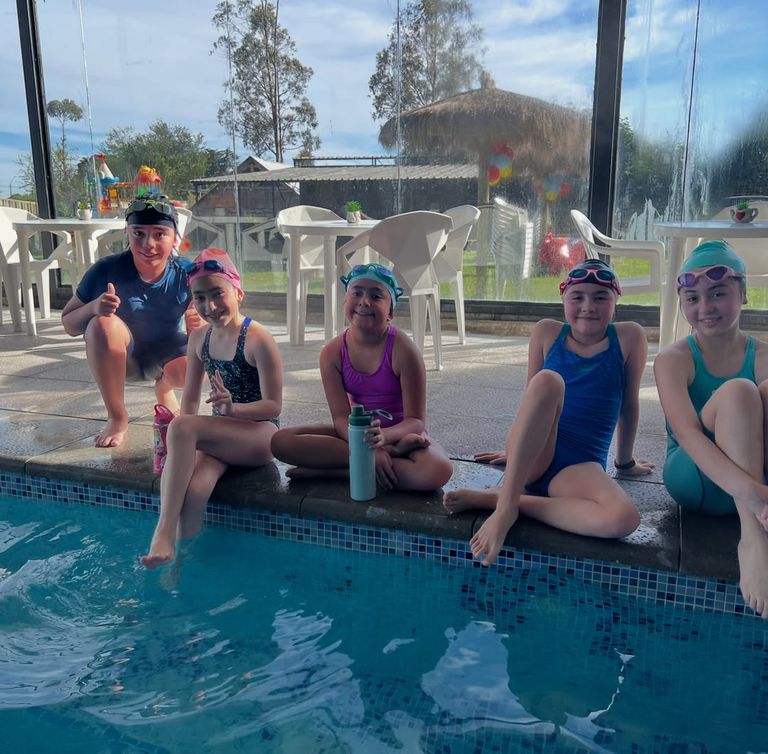 Alumnas y alumnos con gorro y antiparras sentados en el borde de la piscina temperada